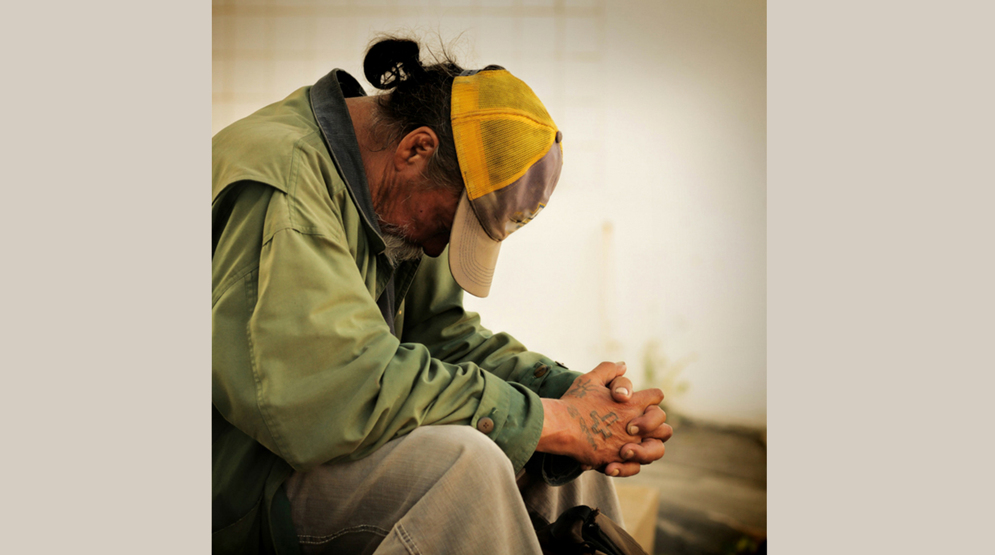 Homeless man wearing baseball cap, sitting head down and hands folded